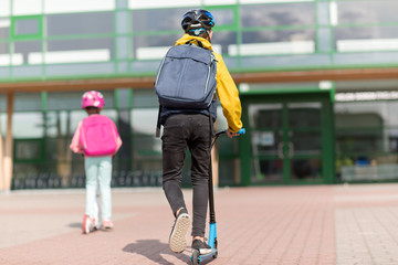 education, childhood and people concept - happy school children with backpacks riding scooters outdoors