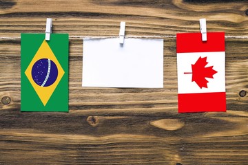 Hanging flags of Brazil and Canada attached to rope with clothes pins with copy space on white note paper on wooden background.Diplomatic relations between countries.