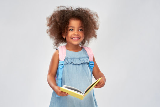 childhood, school and education concept - happy little african american girl with book and backpack over grey background