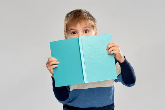 childhood and education concept - little boy hiding behind book over grey background