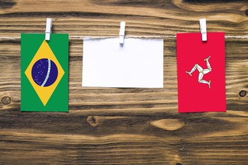 Hanging flags of Brazil and Isle Of Man attached to rope with clothes pins with copy space on white note paper on wooden background.Diplomatic relations between countries.