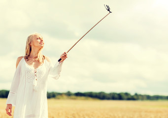 technology, summer holidays, vacation and people concept - smiling young woman in white dress taking picture by smartphone selfie stick on cereal field