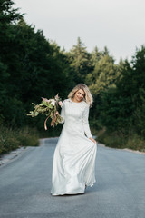 Beautiful elegant bride in lace wedding dress with long full skirt and long sleeves. She is holding a big bouquet of flowers. Outdoors, on the road.