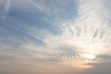 Bright cloudscape. Natural background. White ribbed clouds illuminated by different colors, a trace from an airplane, a luminous spot of the sun.