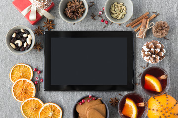 christmas, winter holidays and technology concept - tablet computer, hot mulled wine, dry orange slices and aromatic spices on grey background