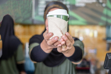 Close up of muslim woman barista wearing hand holding paper cup of coffee and giving to customer. Selective focus on her hand.