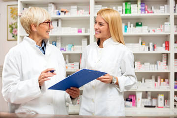 Happy mature and young female pharmacists talking in the pharmacy. Mature woman is holding a clipboard. Healthcare and medicine concept