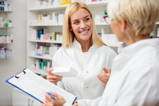 Smiling happy young blond-haired pharmacist holding a box of medications, consulting with her older colleague. Healthcare and medicine concept.