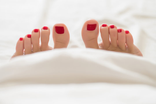 female finger with red pedicure in bed from under the white covers Close-up of covered female feet with red pedicure