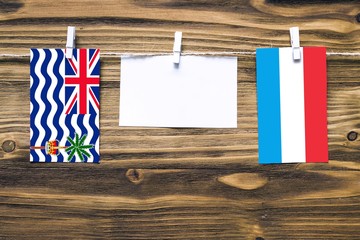 Hanging flags of British Indian Ocean Territory and Luxembourg attached to rope with clothes pins with copy space on white note paper on wooden background.