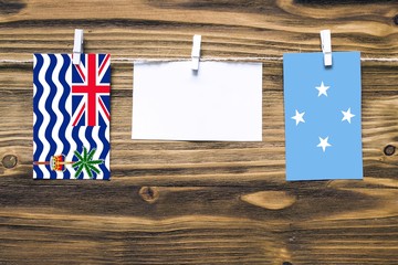 Hanging flags of British Indian Ocean Territory and Micronesia attached to rope with clothes pins with copy space on white note paper on wooden background.