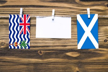 Hanging flags of British Indian Ocean Territory and Scotland attached to rope with clothes pins with copy space on white note paper on wooden background.