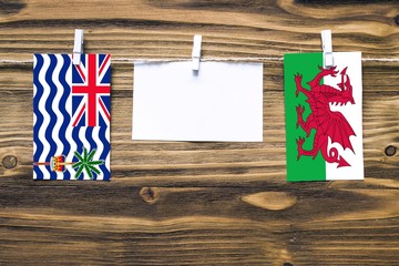 Hanging flags of British Indian Ocean Territory and Wales attached to rope with clothes pins with copy space on white note paper on wooden background.