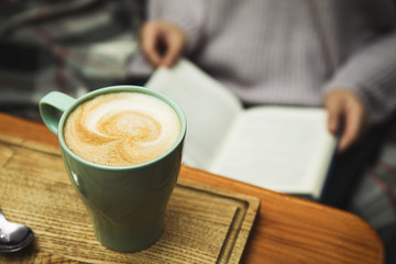 Woman reading book at table, focus on cup of coffee