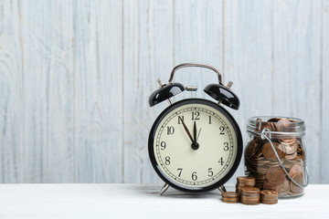 Naklejka premium Black alarm clock, glass jar and coins on white table against wooden background, space for text. Money savings
