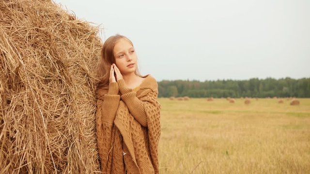 Adorable happy girl posing near haystack in field. Beautiful smiling teenage girl with long hair leaning at hay and looking away in autumn field during harvest