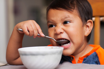 A latino boy taking a bite out of a spoonful of food and sitting unsupported at the dinner table for breakfast.