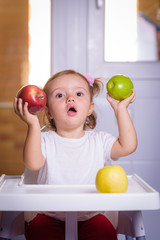 Fototapeta premium Cute baby 1,4 years old sitting on high children chair and eating fruit alone in white and brown kitchen