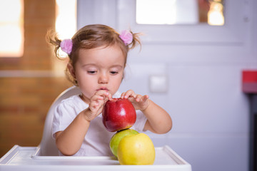 Fototapeta na wymiar Cute baby 1,4 years old sitting on high children chair and eating fruit alone in white and brown kitchen