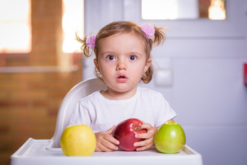 Fototapeta premium Cute baby 1,4 years old sitting on high children chair and eating fruit alone in white and brown kitchen