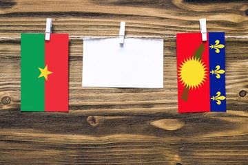 Hanging flags of Burkina Faso and Guadeloupe attached to rope with clothes pins with copy space on white note paper on wooden background.Diplomatic relations between countries.