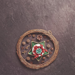 A green fir cone along with red berries is in a white bowl on a green star-shaped plate. Nearby are various decorations (wicker wreath of branches, anise and dried plants). Dark cement background.