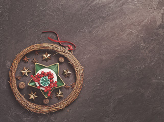 A green fir cone along with red berries is in a white bowl on a green star-shaped plate. Nearby are various decorations (wicker wreath of branches, anise and metal stars). Dark cement background.