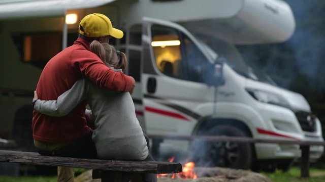 RV Campsite Family Time. Father Hugging His Daughter on Wooden Bench in Front of Campfire and Motorhome.