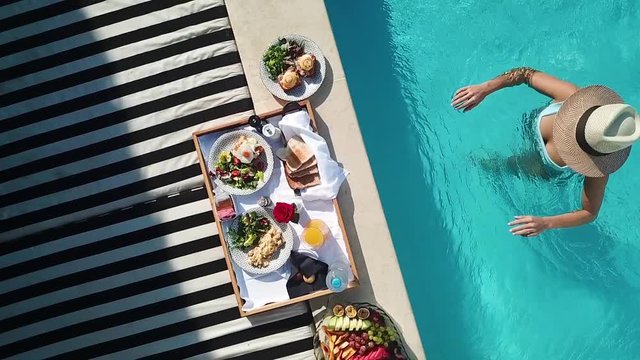 Breakfast for Two Served on Plate on Poolside of Luxury Hotel and Female in Bikini Approaching From Swimming Pool, Bird Eye Slow Motion Aerial View