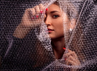 Abstract mysterious portrait girl hidden behind breaking bubble wrap in black background and looking through the gap plastic
