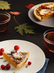 A piece of homemade traditional American pumpkin pie is located on a plate. Close-up. On a black background multi-colored autumn leaves and a cups with tea. Selective focus.