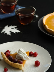 A slice of homemade traditional American pumpkin pie decorated with cream and cranberries is located on a white plate. Close-up. On a black background multi-colored autumn leaves and a cups with tea.