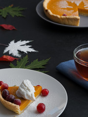 A slice of homemade traditional American pumpkin pie decorated with cream and cranberries is located on a white plate. Close-up. On a black background multi-colored autumn leaves and a cup with tea.