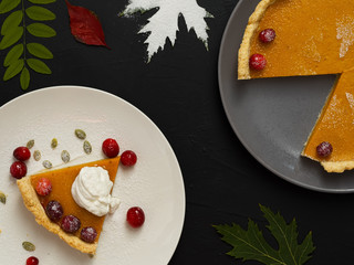 Slice of homemade traditional American pumpkin pie decorated with cream and cranberries on a white plate. Top view. On a black background are colorful autumn leaves and a whole cake on a gray plate.