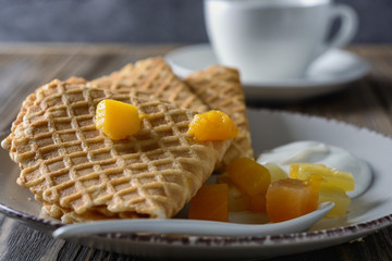 Thin waffles folded as a corner with cream, syrup and slices of fruit on a ceramic plate next to cup of tea on a wooden table ready for breakfast, close up