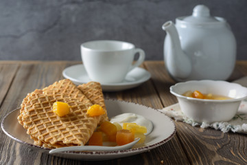 Thin waffles folded as a corner with cream, syrup and slices of fruit on a ceramic plate next to cup and teapot on a wooden table ready for breakfast
