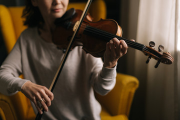 Beautiful young woman musician playing the violin close-up, sitting on soft chair in room with a modern interior. Girl is practicing playing musical instrument at home.