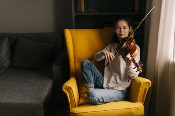 Attractive young woman musician plays the violin sitting on soft chair. Girl is practicing playing musical instrument at home.