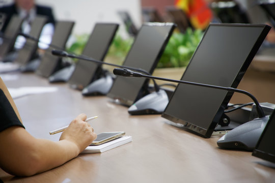 A woman with a smartphone, fountain pen and notebook sits at a table with monitors and intercoms during a conference call. Without a face