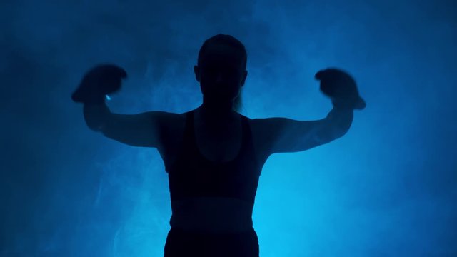 Silhouette of boxer woman showing biceps before sparring on dark background of blue smoke.