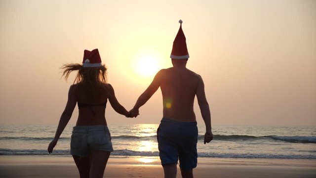 A young couple in red Santa Christmas hats walk into the sea on the beach during a beautiful sunset holding hands. Happy Christmas on holiday at the seaside.