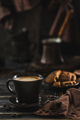A Cup of coffee with foam on a dark wooden background with oatmeal cookies, scattered grains, coffee maker in the background.