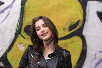 Portrait of european young beautiful smiling woman with dark straight hair in black leather jacket on graffiti wall background