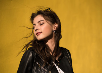 Portrait of european young beautiful smiling woman with dark straight hair in black leather jacket on yellow wall background