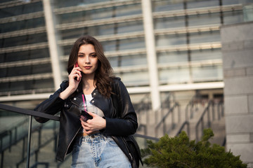 Fototapeta na wymiar Portrait of european young beautiful smiling woman with dark straight hair in black leather jacket and blue jeans talking to phone on city background