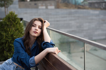 Portrait of european young beautiful smiling woman with dark straight hair in blue shirt and blue jeans sitting on bench