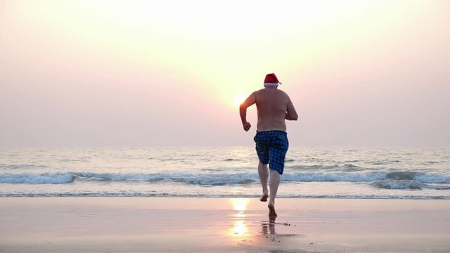 A fat man in a red Santa hat runs funny holding shorts and dives into the sea at sunset. A goofy Christmas image of a running fat Santa Claus in the sea.