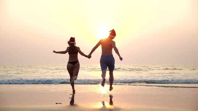 A young couple in red Santa Christmas hats runs into the sea on the beach during a beautiful sunset holding hands. Happy Christmas on holiday at the seaside.