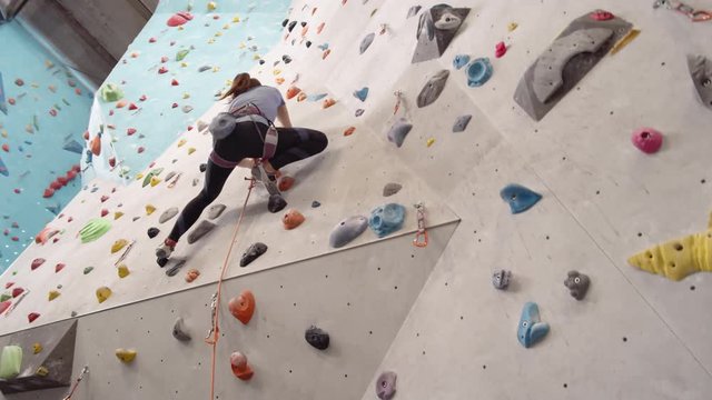 Wide shot of young Caucasian sportswoman wearing safety belay climbing wall in gym