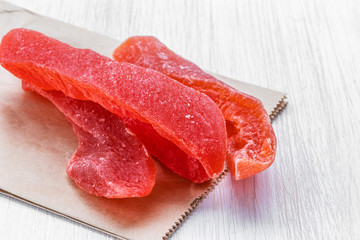 Red dried fruits in sugar lie on a gray paper bag on a white wooden background, side view from above